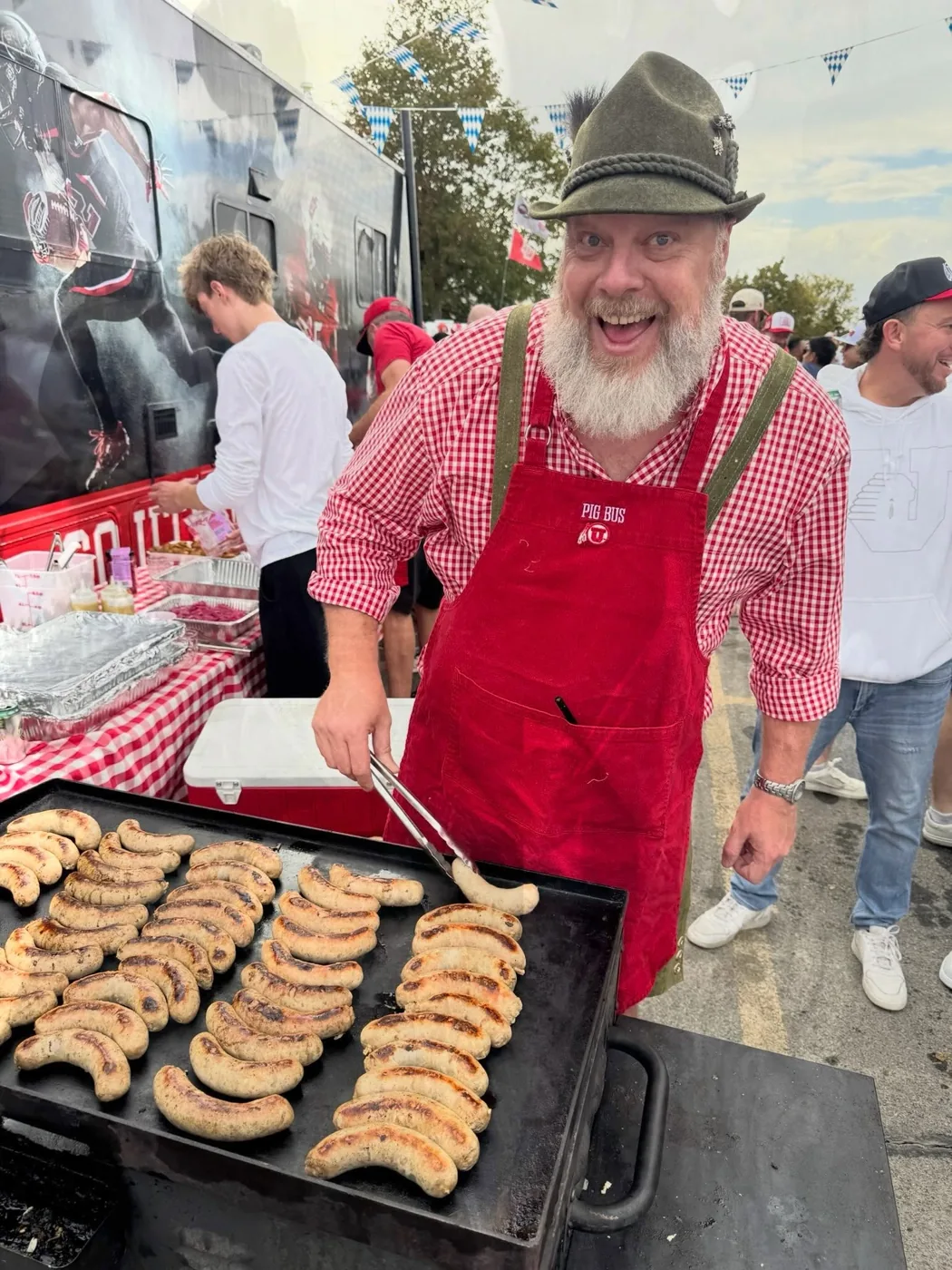 A crew member in an Alpine hat and Pig Bus apron working a griddle of brats