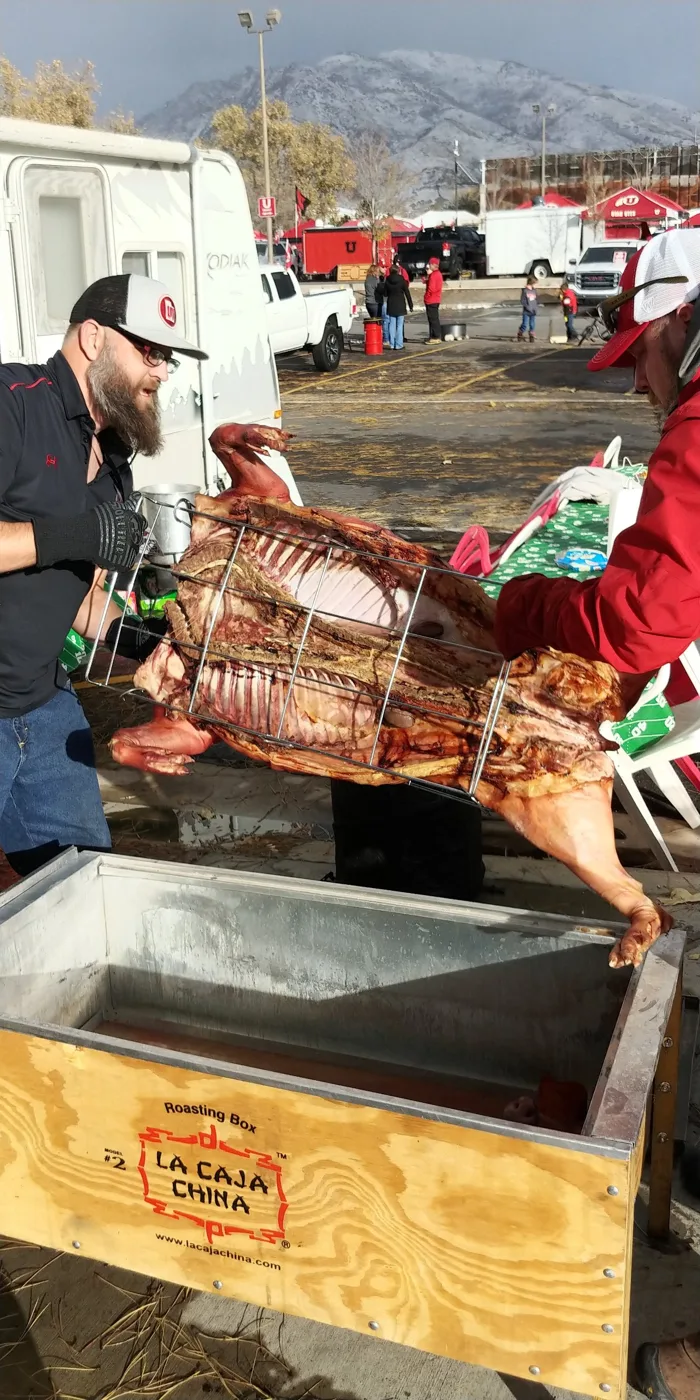Two crew members lifting a roasted whole pig in its rack out of the roasting box, snowy mountains behind